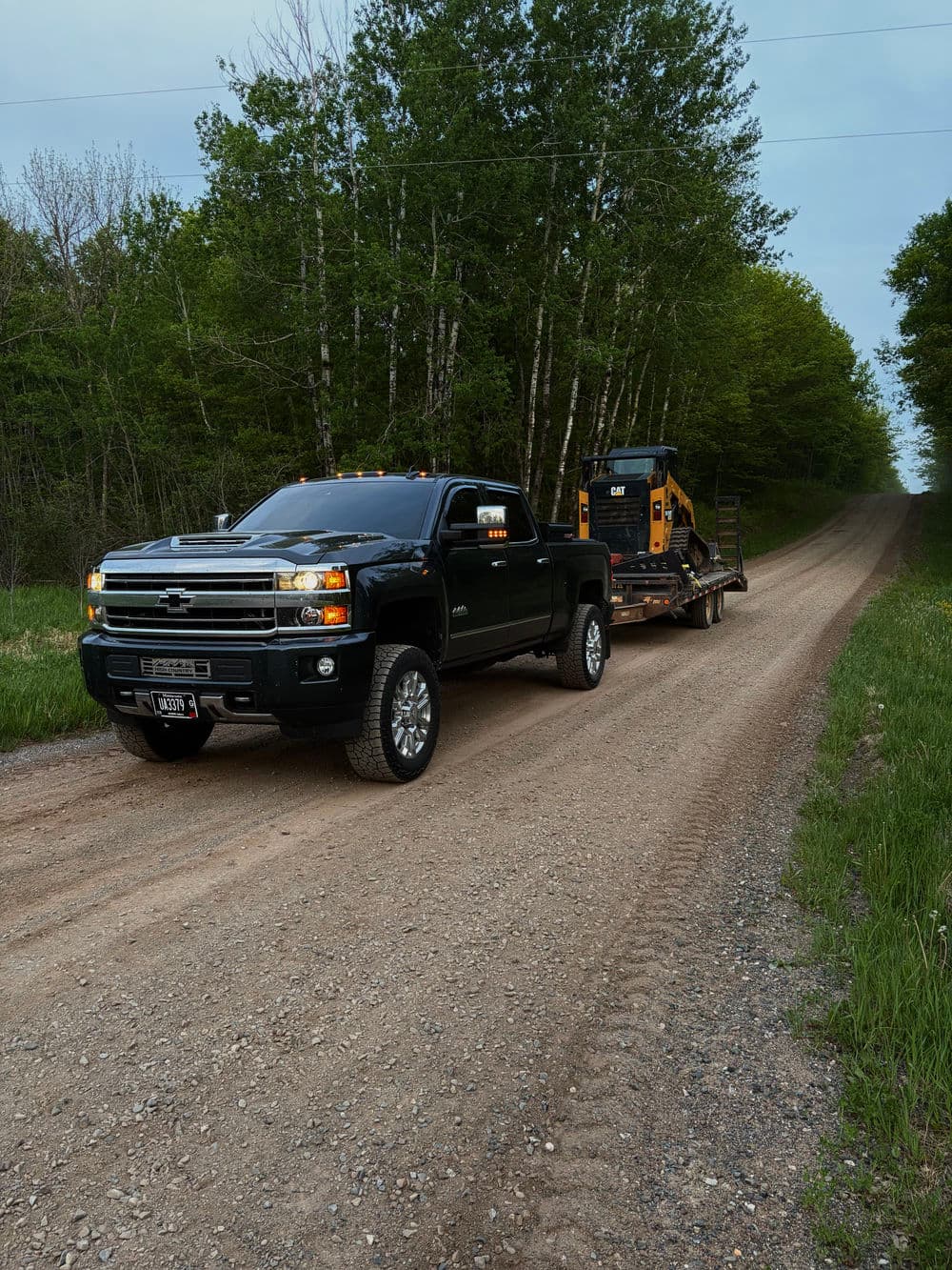Black Chevrolet truck hauling heavy equipment on a gravel road surrounded by trees.